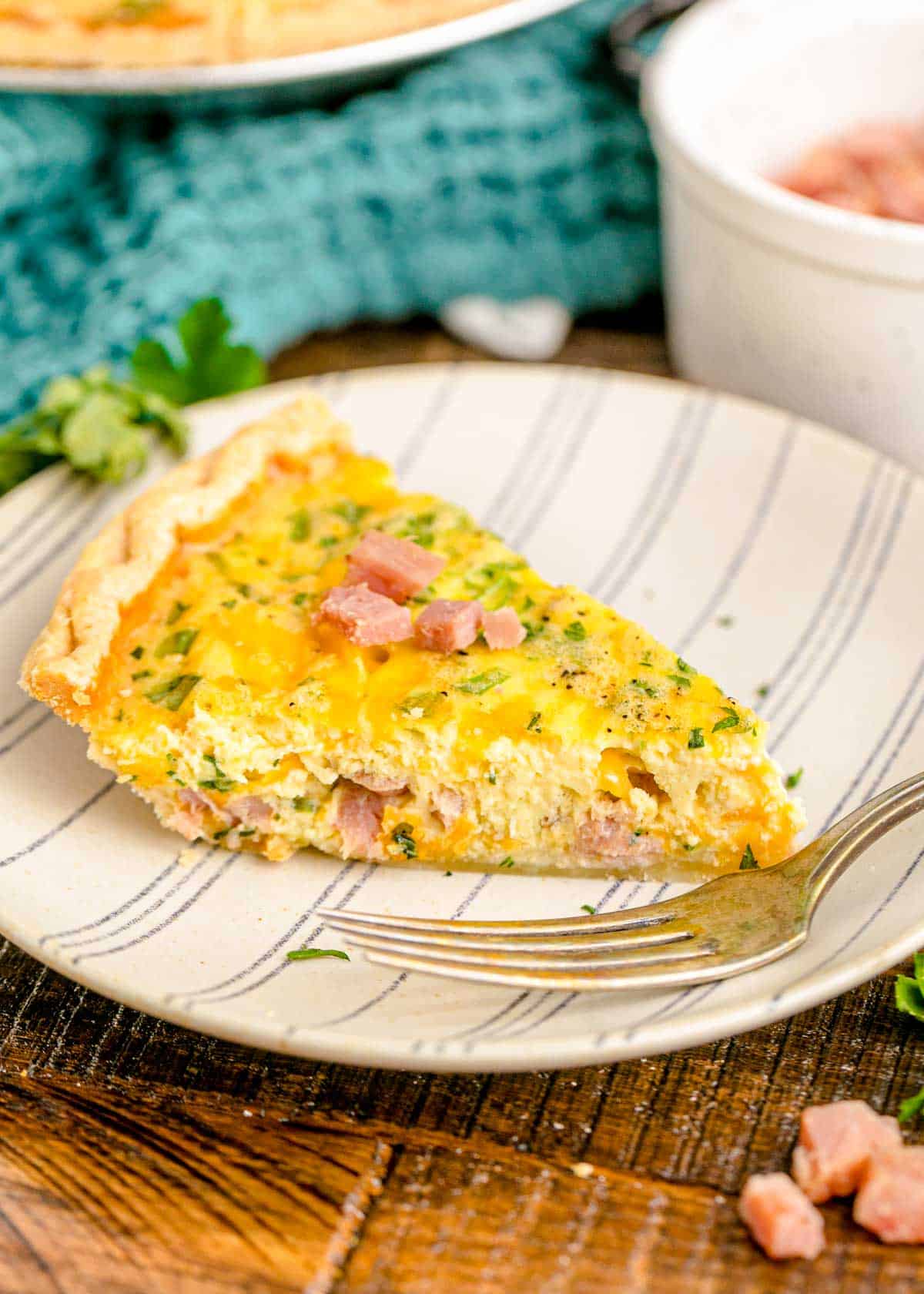 A slice of ham and cheese quiche on a white and striped plate with a fork.