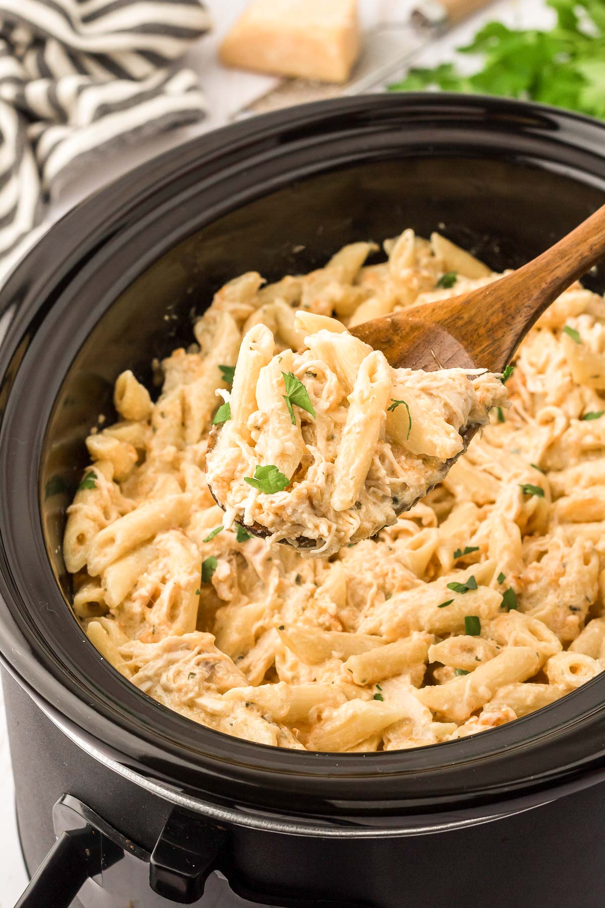 A wooden spoon scooping garlic chicken pasta out of a crockpot.