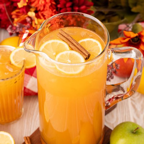 A pitcher of apple lemonade on a wooden table with apples, cinnamon sticks, and lemons around it.